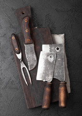 Vintage meat knife and fork and hatchets with vintage chopping board and black table background. Butcher utensils. Close up