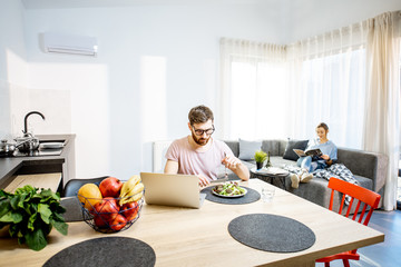 Man working with laptop while eating healthy salad with woman on the background resting on the couch in the modern studio