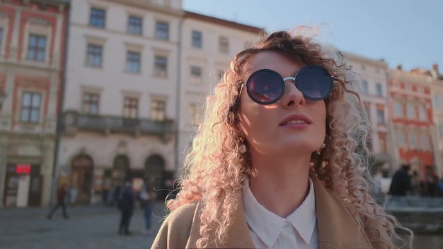 Portrait of gorgeous smiling curly woman in stylish look confidently walks down the street, weared sunglasses. Fashion blogger with golden curls standing on street in sunlight. Portrait, urban