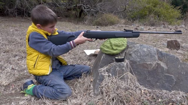 Young boy pulls the trigger on small air rifle leaning against a rock in slow motion