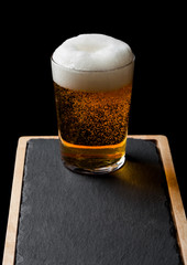 Glass of lager beer with foam and bubbles on stone board on black background.