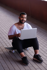 Smiling African-American guy is sitting on a bridge sidewalk, holding the laptop on his knees and looking at a camera