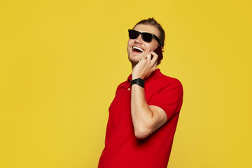 Joyful businessman wearing red polo shirt and sunglasses having phone call on yellow background. Confident guy talking on smartphone while standing on yellow backdrop