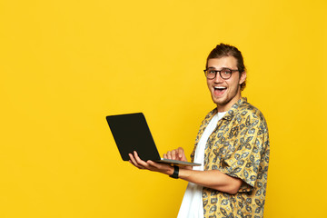 Attractive young hipster boy in eyeglasses holding laptop computer and laughing joyful isolated over yellow background.