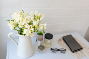 Close up of white freesias in jug on dressing table with bottles, scarf, purse and glasses in background (selective focus)