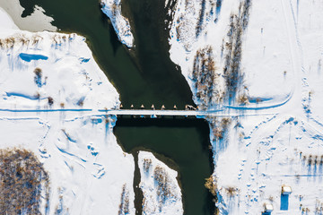 Aerial top view of a bridge over Sura River, white snowy land and road, trees and country houses on a sunny winter day. Bessonovka, Penza oblast, Russia