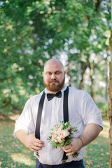 Stylish bearded fat groom with wedding bouquet in the park.