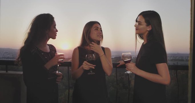 Beautiful three ladies in the top of building at the balcony have a girls party they drink some glass wine and have a nice conversation background amazing sunset view.