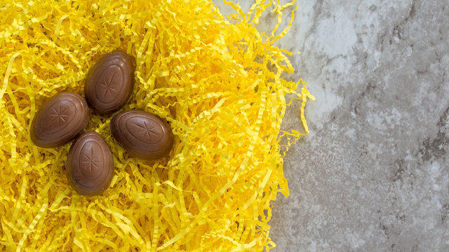 four chocolate Easter eggs nestled in yellow Easter grass on a gray marble counter with copy space