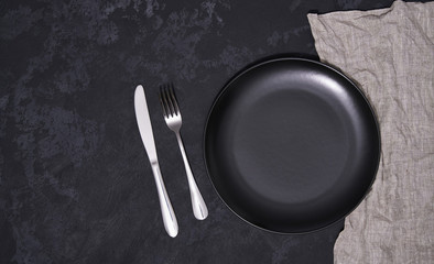Empty black plate and cutlery on a black background, top view.