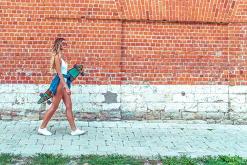 Beautiful blond woman with long hair walks brick wall, summer in city. White bodysuit with jeans, longboard board. Free space for text. Concept girl hipster goes event weekend, walk to meeting.