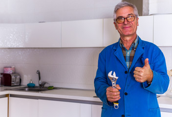 Senior handyman in blue uniform shows thumbs up while holding wrench