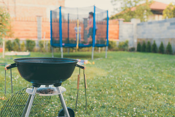 Kettle Charcoal BBQ Barbecue Grill in garden or backyard. Blurred Outdoor trampoline in the background. Family Home Backyard BBQ Party Or Picnic Conceptual Scene.