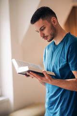 Student reading a book while standing and walking in the room next to the windows.