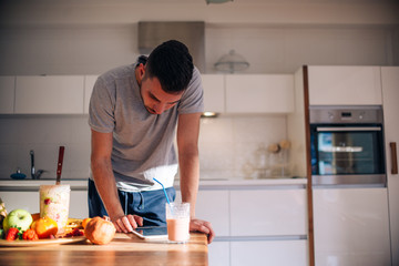 Vegan man in his 20's has a morning preparation of a smoothie while looking at a recipe on his tablet.