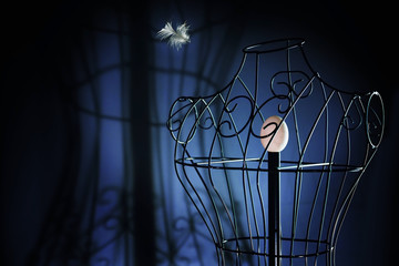 egg in a metal tailor doll like in a bird cage and a flying feather, surreal art concept for longing and loneliness, dark blue background with copy space