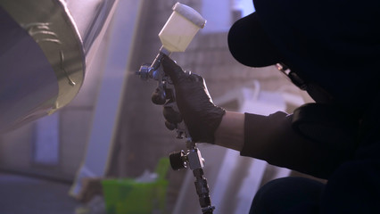 A professional guy (man) in the garage covers the yacht (transport) with a mini-jet (sprays) by applying paint (ceramics) to the surface using a protective suit dispenser and protective glasses.