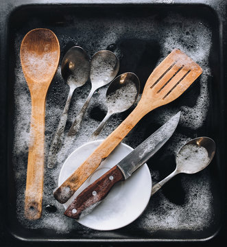 Washing dishes. A plate, a knife, wooden kitchen spatulas and spoons in the detergent foam on a black oven-tray. Top view. Guest worker concept.