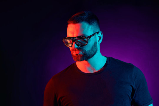 Close up portrait of a bearded serious man in styled clubber glasses at studio. High Fashion male model in colorful bright neon lights posing on black background.Art design concept