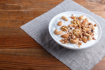 White plate with greek yogurt, granola, almond, cashew, walnuts  on brown wooden background.