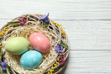Wicker nest with painted Easter eggs and flowers on wooden table, top view. Space for text