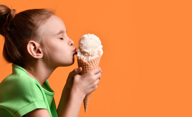 Little baby girl kid kissing vanilla ice cream in waffles cone on yellow orange background in green t-shirt - 252967963