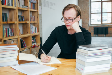A young man in a black sweatshirt and glasses, a student or teacher, reads a book in the library, and writes information into a notebook. studies information, prepares for exams.