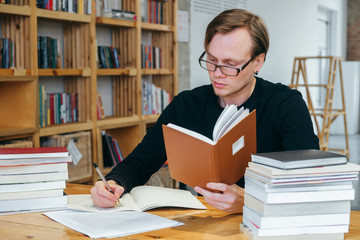 A young man in a black sweatshirt and glasses, a student or teacher, reads a book in the library, and writes information into a notebook. studies information, prepares for exams.