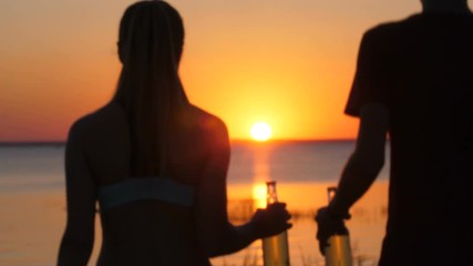 Silhouette of couple with beer bottles walking towards setting sun on beach while spending summer vacations at seaside. Couple of travelers with beers in hands going to rest on shore in twilight - Powered by Adobe