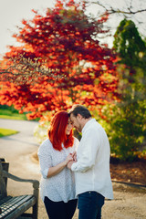 Naklejka premium beautiful and stylish redhead woman in a white blouse standing in a sunny summer park with her handsome husband near flower trees