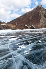 Lake Baikal in March. The view from the beautiful ice with cracks on the 130 km of the Circum-Baikal railway with the stone arch bridge across the Shabartuy River, bordered by rocky mountains