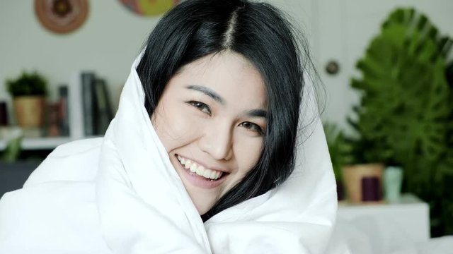 Young beautiful asian woman laying in bed under blanker and jump up to camera, close up shot. Surprise morning concept.