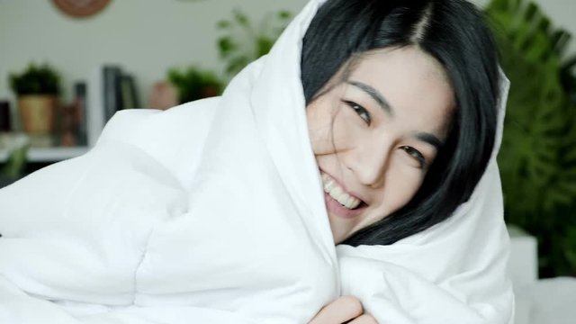 Young beautiful asian woman laying in bed under blanker and jump up to camera, portrait. Surprise morning concept.