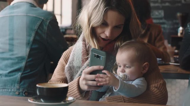 young mother with baby in cafe using smartphone drinking coffee relaxing in busy restaurant enjoying motherhood