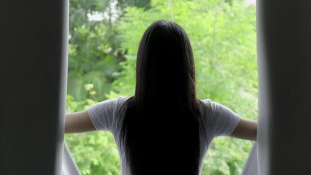 Slow motion of young beautiful asian woman open bedroom curtain to see green trees background. Relaxing home concept.