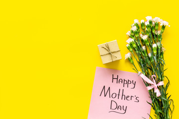 Happy Mother's Day concept. Hand lettering near bouquet of pink carnation and gift box on yellow background top view space for text