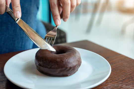Closeup image of woman's hands cutting a piece of chocolate donut by knife and fork for breakfast on wooden table