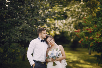 A young and beautiful bride and her husband is standing in a summer park with bouquet of flowers