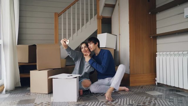 Young man and woman are taking selfie with smartphone making heart shape with fingers sitting on floor of new apartment with boxes in background. Relocation and fun concept.