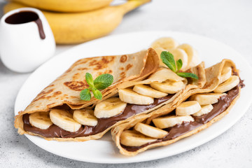Crepes stuffed with chocolate spread and banana on white plate. Thin pancakes, blini. Sweet dessert. - obrazy, fototapety, plakaty