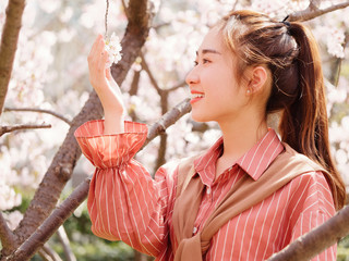 Outdoor portrait of beautiful young Chinese girl in red shirt posing among pink blossom cherry tree brunch in spring garden, beauty, summer, emotion, expression and people concept. 