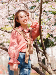 Outdoor portrait of beautiful young Chinese girl in red shirt posing among pink blossom cherry tree brunch in spring garden, beauty, summer, emotion, expression and people concept. 