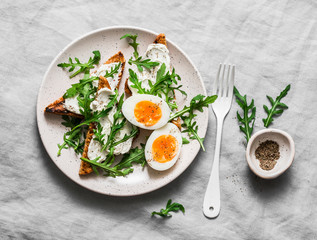 Delicious breakfast, brunch or snack - cream cheese toast, arugula salad and soft boiled egg on a light background, top view