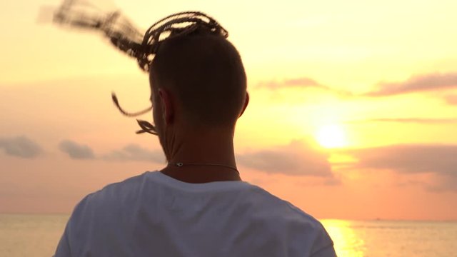 Young guy dancing sunset time and turns his head with dreadlocks on the tropical beach near sea water. Close up portrait. Happy handsome man with dreadlocks turns his head, slow motion