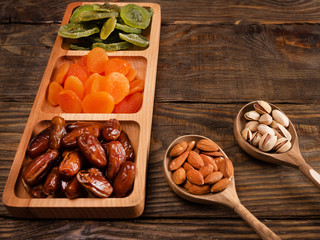 Dates, dried apricots and kiwis in a Compartmental dish and nuts in a wooden spoon on a dark wooden table.