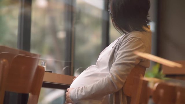Side view of Young and beautiful Chinese pregnant women sitting in the cafe the window, stretching lazily, and touching their stomachs, pregnant Asian businesswoman lifestyle 4k footage