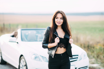 A beautiful woman dressed in trousers and a leather jacket waiting for a white cabriolet car