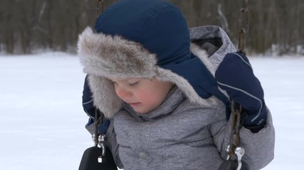 kid swaying on swing. Cute little boy (toddler, two year old) in hat with fur trim and earflaps.  Happy child. Winter, cold weather, outdoor