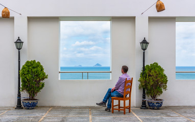 A man looks from the balcony at the sea on a Sunny day. Decorative plant and an old lantern near the white wall of the terrace.