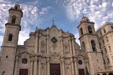 Fototapeta na wymiar Facade of cathedral in Havana, Cuba with sandstone towers and brass bells and cross symbols on roof under bright blue sky and white clouds